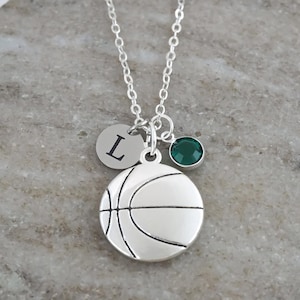 May include: A silver-toned necklace featuring a basketball charm, a small circular disc charm engraved with the letter L, and a round dark green crystal charm, all suspended from a delicate silver-toned chain link necklace.