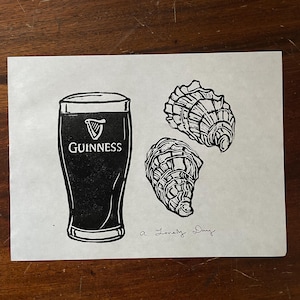 Pu&ograve; includere: Una stampa in linoleum in bianco e nero di un bicchiere di birra Guinness e tre ostriche. Il testo "a lovely day" &egrave; scritto sotto l'immagine.