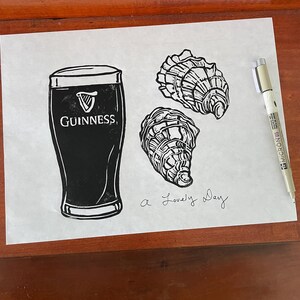 Pu&ograve; includere: Un'illustrazione in bianco e nero di una pinta di birra Guinness e due ostriche. Il testo "a lovely day" &egrave; scritto sotto l'immagine.
