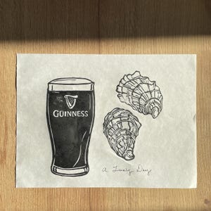 Pu&ograve; includere: Un'illustrazione in bianco e nero di una pinta di Guinness, due ostriche e il testo "a lovely day".