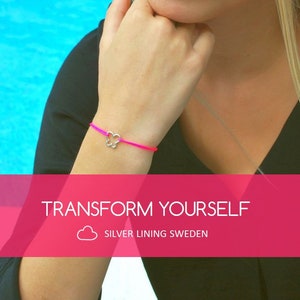 Può includere: Un braccialetto di corda rosa e viola con un ciondolo a forma di farfalla argentata. Il braccialetto è indossato al polso di una persona. Il testo "TRANSFORM YOURSELF" e "SILVER LINING SWEDEN" è visualizzato su uno sfondo rosa.