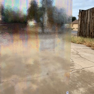 May include: A clear iridescent glass sheet with a textured surface. The glass has a rainbow sheen and is held up in front of a blurry background.