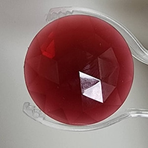 May include: A close-up of a red, faceted, glass gem held in a clear plastic clamp.