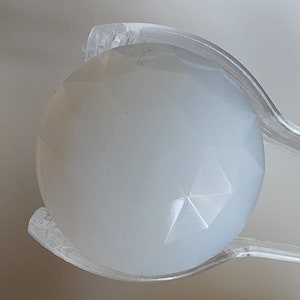 May include: A white, faceted, round gemstone held in clear plastic tweezers.