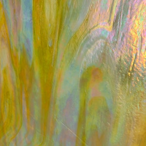 May include: Close-up of a sheet of iridescent stained glass. The glass has a swirling pattern of yellow, gold, and hints of blue and pink. The surface reflects light, creating a shimmering effect. Ideal for craft projects.