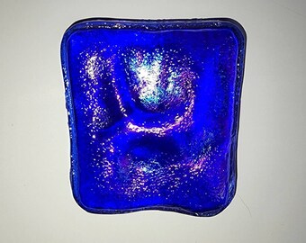 May include: A square blue glass tile with an iridescent finish. The tile has a textured surface and a slightly wavy edge.