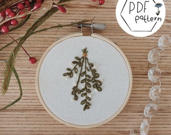 May include: A white embroidery hoop with a green and white embroidered sprig of mistletoe. The hoop is on a wooden surface with a red berry garland and a white hexagonal sign that says "PDF pattern".