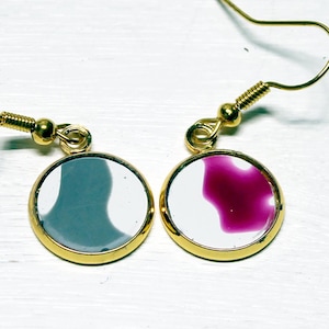 May include: A pair of dangle earrings featuring round gold-toned metal settings. One earring contains a white inlay with a gray abstract shape, while the other contains a white inlay with a magenta abstract shape. Both have gold-toned ear wires.