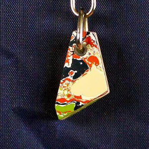 May include: A colorful pendant with a unique, abstract design. The pendant features a mix of red, black, white, and green hues, with a gold-colored metal border and a silver-colored clasp. The pendant is triangular in shape.