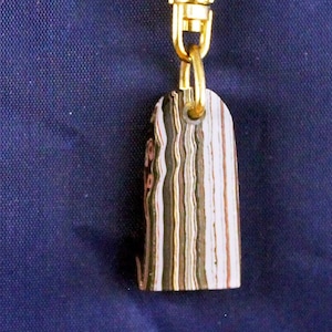 May include: A rectangular keychain pendant with a gold-colored clasp. The pendant features a layered pattern of brown, white, and pink tones. The keychain is hanging against a dark blue background.