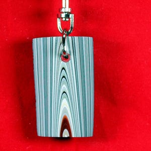 以下が含まれることがあります： グレー、白、茶色のストライプ模様の長方形のキーホルダーペンダント。銀色の金属製の留め金が付いています。背景は無地の赤色です。