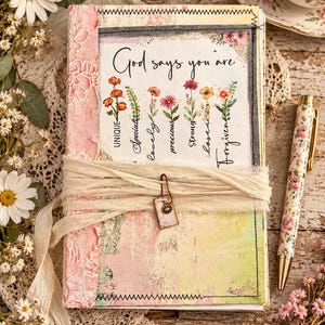 May include: A journal with a pink and yellow distressed cover featuring the text "God says you are" followed by a list of words including "UNIQUE," "Special," "lovely," "precious," "Strong," "chosen," and "Forgiven" alongside floral illustrations.
