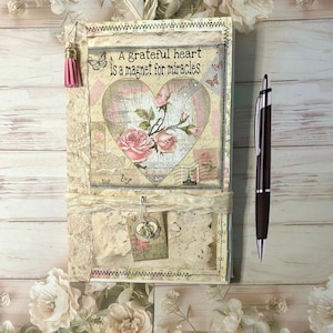 May include: A handmade journal with a heart-shaped design featuring pink roses and the text "A grateful heart is a magnet for miracles." The journal has a pink tassel and a pen.