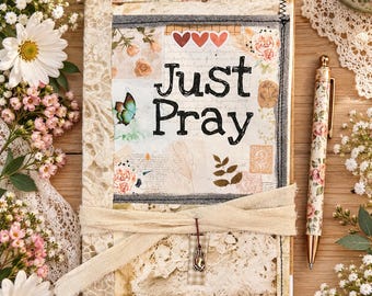 May include: A rectangular journal with a cream-colored lace cover, featuring a central collage with the text "Just Pray" in black lettering, three small red hearts, a butterfly illustration, floral motifs, and a dangling metal charm of praying hands.