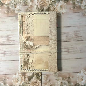 May include: A vintage-style journal with a cream-coloured cover adorned with lace, paper, and ribbon accents. The journal is bound with a cream-coloured twine and features a decorative edge stitching. The overall aesthetic is soft and romantic.