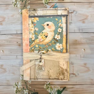 May include: A vintage-style journal with a bird illustration on the cover. The cover features a cream-colored bird with floral accents perched on a branch against a teal background. The journal is bound with a cream-colored ribbon and a small tag.