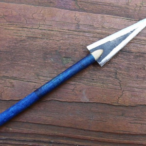 Könnte beinhalten: Ein handgefertigter Miniatur-Speer mit silberfarbener Pfeilspitze und einem blau lackierten Holzschaft. Der Speerkopf hat schwarze Akzente. Der Schaft ist ein Bleistift mit einem blau lackierten Abschnitt.