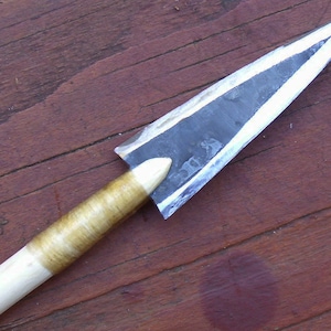 May include: A close-up of a hand-forged arrowhead with a wooden shaft. The arrowhead is made of metal and has a sharp point. The shaft is wrapped in a brown material.