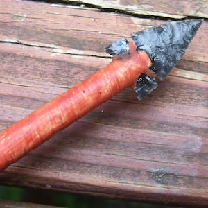 May include: A red and black arrowhead with a wooden shaft. The arrowhead is made of flint and has a sharp point. The shaft is made of wood and is about 15 cm long.