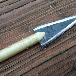 May include: A close-up of a wooden arrow with a metal arrowhead. The arrowhead is sharp and pointed, and the shaft is wrapped in a light brown material.
