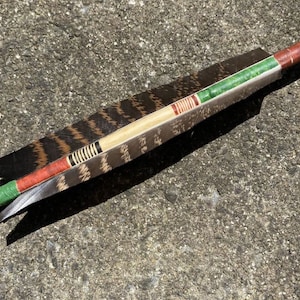 May include: A close-up of a brown and black fletched arrow with red, green and white painted bands. The arrow is lying on a grey concrete surface.