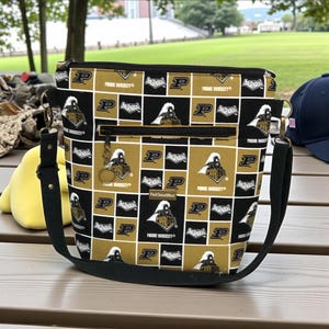 Op de afbeelding: Zwarte en gouden crossbodytas met een herhaald patroon van het Purdue University logo en tekst. De tas heeft een zwarte verstelbare schouderriem, een voorvak met rits en een ritssluiting aan de bovenkant. Gemaakt door ParkStreetWest.