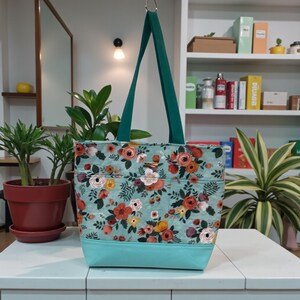 May include: A floral tote bag with a light blue base and handles. The bag features a pattern of red, orange, and white flowers on a light blue background. The handles are a dark green color.