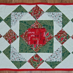 May include: A white table runner with a red, green, and white patchwork design. The runner features a repeating pattern of diamond shapes with a central square containing the words "Peace on Earth" and "Deck the Halls".