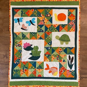 May include: A white quilt with a green border and orange and green patchwork squares. The quilt features appliqued images of a snail, turtle, dragonfly, fish, and lily pads.