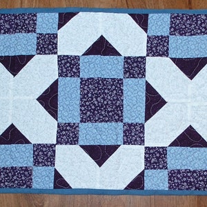 May include: A white and blue quilted table runner with a geometric pattern. The runner is made of fabric squares sewn together in a repeating pattern.