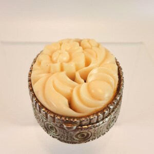 May include: An oval-shaped trinket box featuring a cream-colored, carved floral relief lid set into a textured, metallic silver-toned base with circular embossed patterns along the side. The lid displays a detailed sculpted flower design.