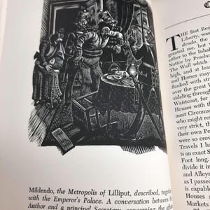 K&ouml;nnte beinhalten: Schwarz-Wei&szlig;-Illustration aus einem Buch, das eine Szene mit mehreren Figuren in historischer Kleidung zeigt, die sich um einen Tisch versammelt haben. Der Text unter dem Bild lautet "Mildendo, die Metropole von Lilliput..." und beschreibt ein Gespr&auml;ch.