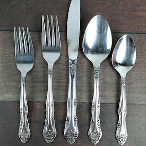 May include: A set of five silver-colored vintage silverware pieces, including two forks, a knife, and two spoons. The handles feature an ornate floral design. The silverware is arranged on a dark wooden surface.