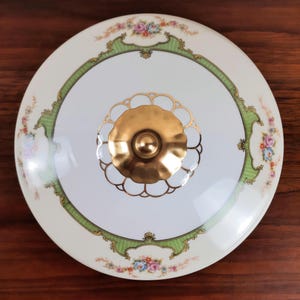 May include: A round, white ceramic lid with a gold handle. The lid features a green and gold border design with floral accents. The handle is a gold-colored, decorative knob. The overall design is vintage and elegant.