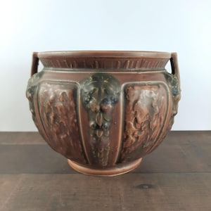 May include: A brown ceramic planter with a wide, rounded body and two small handles. The planter features detailed relief carvings of grape vines and foliage. The rim and base are a slightly lighter shade of brown.