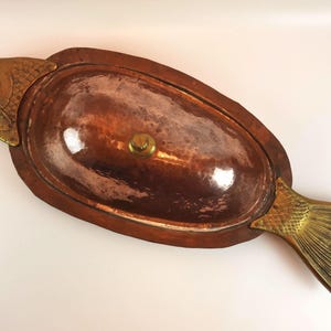 May include: An oval copper serving dish with a hammered texture and a central brass knob on the lid. The dish features decorative brass fish head and tail attachments at opposite ends, creating a cohesive fish-shaped design for the vessel.