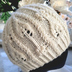 May include: A white knitted hat with a cable knit pattern. The hat has a textured surface and a slightly slouchy fit.