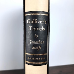 K&ouml;nnte beinhalten: Ein gebundenes Buch mit dem Titel "Gullivers Reisen" von Jonathan Swift. Der Buchr&uuml;cken hat ein schwarzes Etikett mit goldfarbenen Buchstaben und dekorativen goldenen Verzierungen. Das Buch hat einen cremefarbenen Einband und das Wort "HERITAGE" ist unten auf dem Etikett aufgedruckt.