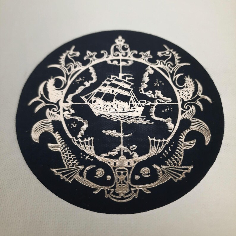K&ouml;nnte beinhalten: Ein kreisf&ouml;rmiges, marineblaues Emblem mit einem detaillierten silbernen Design. Das Design zeigt ein Segelschiff, Fische und kunstvolle Verzierungen. Das Emblem ist wahrscheinlich ein Aufn&auml;her oder ein dekoratives Element.