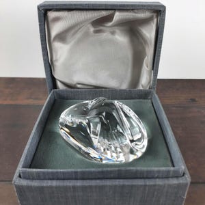 May include: A clear, sculpted glass paperweight in a gray presentation box. The paperweight has a rounded, organic shape with intricate internal facets. The box is lined with gray satin and rests on a dark wooden surface.