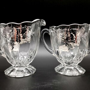 May include: A set of two clear glass sugar and creamer with silver floral designs. The creamer has a single handle and the sugar bowl has two handles.