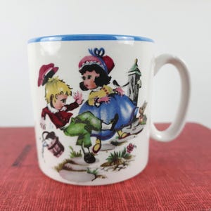 May include: White ceramic mug with a blue rim and a curved handle. The mug features a colorful illustration of two children in vintage clothing, with a small bucket and a building in the background. The artwork has a storybook style.