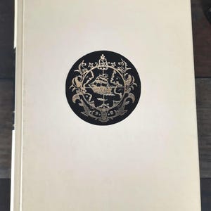 K&ouml;nnte beinhalten: Ein cremefarbenes Hardcover-Buch. Auf dem Einband befindet sich ein schwarzes rundes Emblem mit einer goldenen Schiffillustration, umgeben von aufwendigen goldenen Details. Der Buchr&uuml;cken ist auf der linken Seite sichtbar.