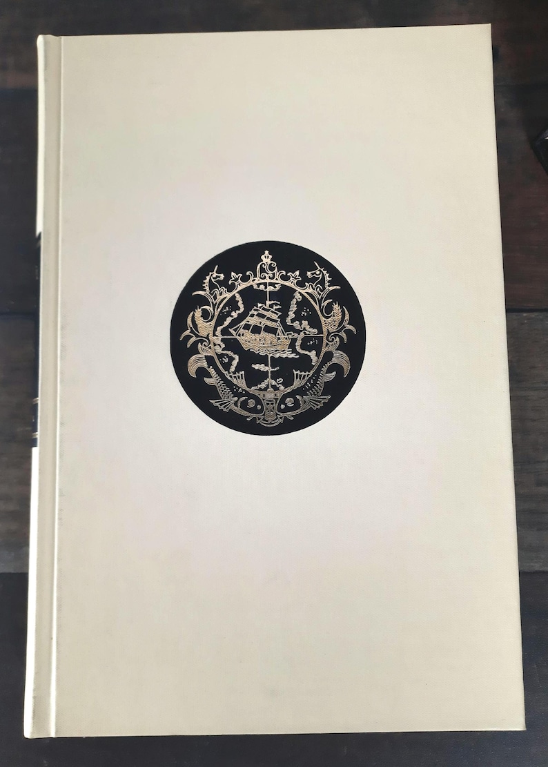 K&ouml;nnte beinhalten: Ein cremefarbenes Hardcover-Buch. Auf dem Einband befindet sich ein schwarzes rundes Emblem mit einer goldenen Schiffillustration, umgeben von aufwendigen goldenen Details. Der Buchr&uuml;cken ist auf der linken Seite sichtbar.
