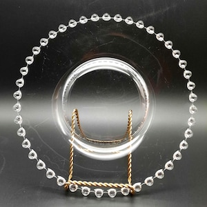 May include: A clear glass charger plate with a beaded edge. The plate is on a gold stand.