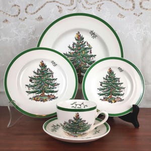 May include: A white Christmas dinnerware set with a green trim. The set includes plates of various sizes, a teacup, and a saucer. Each piece features a decorated Christmas tree design.