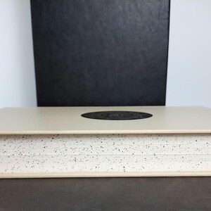 K&ouml;nnte beinhalten: Ein beiges Buch mit gesprenkeltem Rand und einem dunklen kreisf&ouml;rmigen Design auf dem Einband. Eine schwarze Box steht hinter dem Buch. Das Buch liegt auf einer dunklen Oberfl&auml;che.