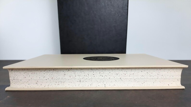 K&ouml;nnte beinhalten: Ein beiges Buch mit gesprenkeltem Rand und einem dunklen kreisf&ouml;rmigen Design auf dem Einband. Eine schwarze Box steht hinter dem Buch. Das Buch liegt auf einer dunklen Oberfl&auml;che.