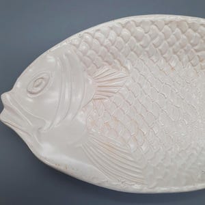 May include: White ceramic fish-shaped serving platter. The platter features a detailed embossed design of a fish, including scales, fins, and facial features. The serving dish is ideal for seafood or decorative purposes.