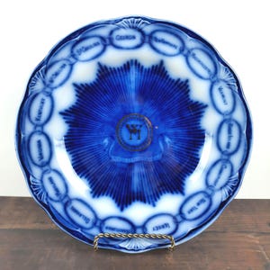 May include: A blue and white antique plate with a scalloped edge. The plate features a central blue starburst design and is encircled by the names of U.S. states. A gold emblem with the letters "W" and "M" is in the center.